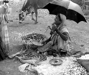 a woman sits on the ground under an umbrella, selling vegetables