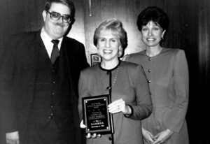 a woman holds an award and smiles. on each side of her is another smiling man and woman. 