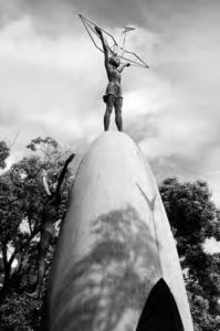 a figure of a girl stands above a structure that is a cross between an egg/ dome, and arch. the girl stands tall with her arms outstretched above her head, holding a metal paper crane. 