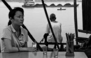 a woman sits at a bar while a man squats and looks out at the water